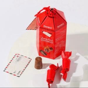 صندوق شوكولاتة شوكوميل أحمر سداسي الشكل، بتصميم مستوحى من البريد، مع بطاقة بريدية فارغة وشوكولاتة حليب مع كورن فليكس (رقائق الذرة) ظاهرة جزئياً وقطعتين مغلفة باللون الأحمر على طاولة بيضاء