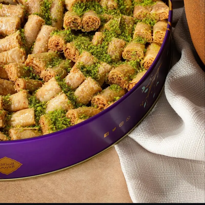 أصابع بقلاوه بالفستق المذاق العربي أصابع بقلاوة ملفوفة مغطاة بالفستق الحلبي في علبة دائرية بنفسجية.